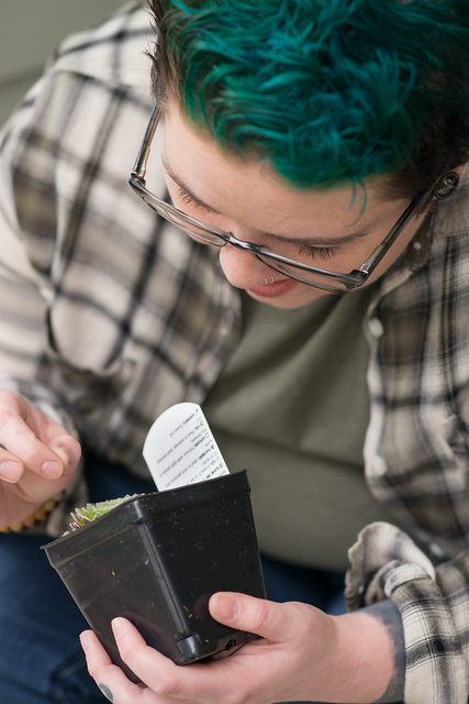 Person wearing glasses examines a potted seedling in the outdoors.