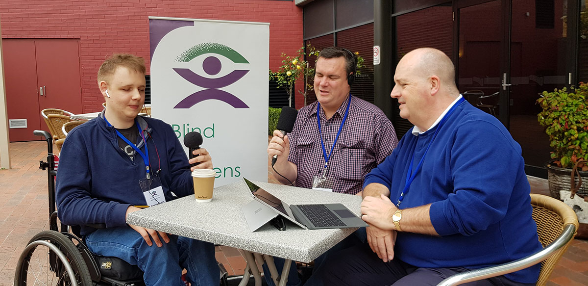 Three people participate in a radio interview at an outdoor table, a BCA banner is visible