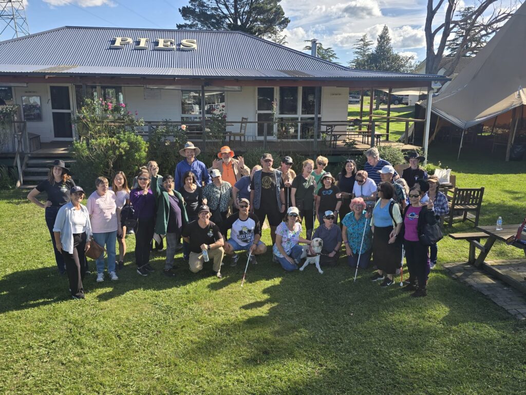 A group of 30 people, including BCA members, volunteers and support workers, standing together outside the entrance to a farm café at Snake Creek Farm Club. The group is arranged closely together, smiling and facing the camera.